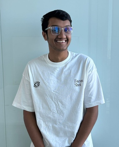 A young man in a white T-shirt and blue sunglasses smiles in front of a clear glass background.