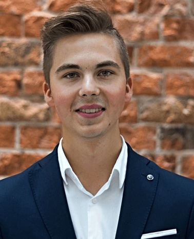 A young man in a dark suit and white shirt smiling in front of a brick wall.