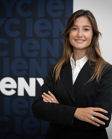 A confident woman in a professional suit stands with her arms crossed, smiling in front of a company logo.