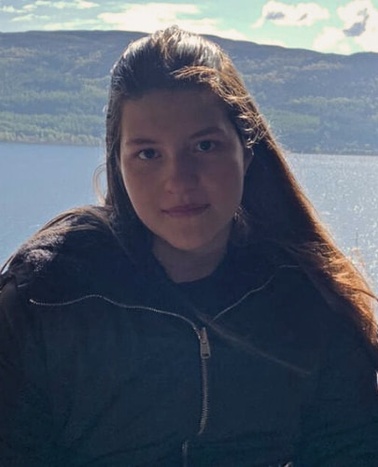 A woman with long brown hair, wearing a black jacket, sitting in front of a scenic lake view.