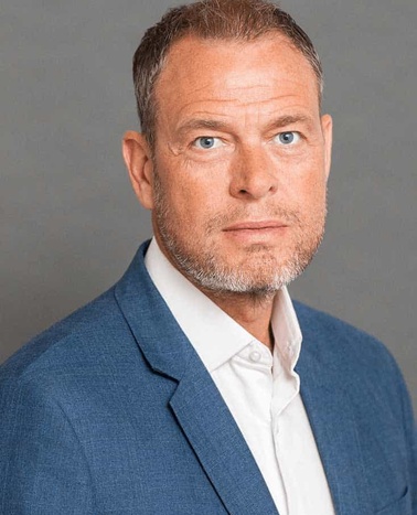 A portrait of a middle-aged man with light stubble, looking directly at the camera, wearing a blue blazer over a white shirt.