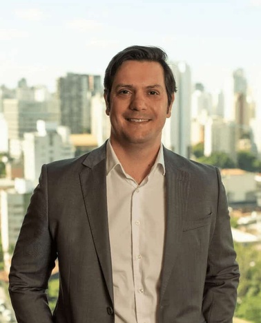 A man in a business suit smiling in an office environment with a cityscape background.