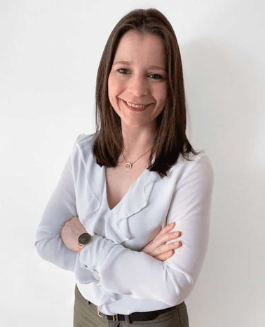 A woman with a friendly smile, wearing a light blue blouse and standing with her arms crossed.