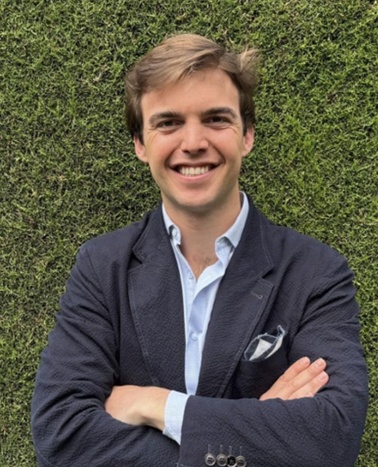 A smiling young man with short hair, wearing a blazer and standing against a green backdrop.