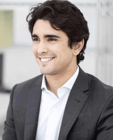A smiling young man with dark curly hair wearing a gray suit and white shirt in an office environment.