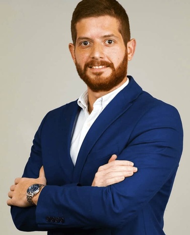 A man in a blue blazer and white shirt standing with his arms crossed, sporting a beard and smiling at the camera.