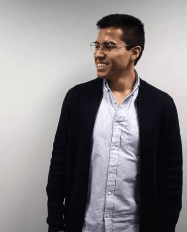 A smiling man wearing glasses, a blue shirt under a black jacket, standing against a plain background.