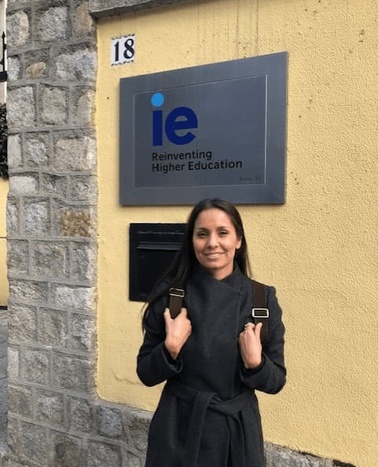 A woman in a black coat smiling in front of a building with a sign that reads 'ie Reinventing Higher Education'.