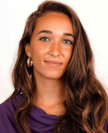 A portrait of a young woman with long brown hair, wearing a purple blouse and hoop earrings, against a plain white background.