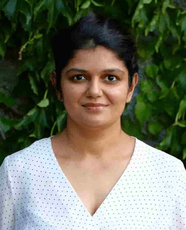 A portrait of a woman in a white polka dot blouse standing in front of a leafy background.
