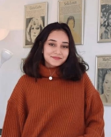 A young woman wearing an orange sweater standing in a room with framed pictures on the wall behind her.