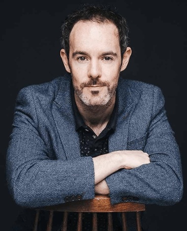 Portrait of a middle-aged man with a beard, resting his arms on the back of a chair, looking directly at the camera against a dark background.