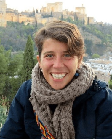 A smiling woman with short hair wearing a scarf, with the Alhambra in the background.