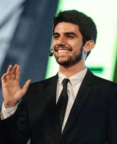 A young man in a suit is smiling and waving, with a microphone headset on, standing on a stage.