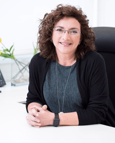 A professional middle-aged woman with curly hair smiling while sitting at a desk in an office.