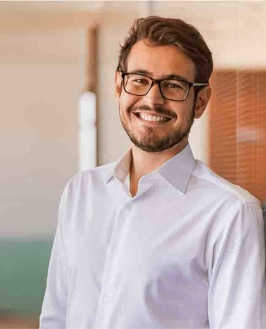 A smiling man wearing glasses and a white shirt standing indoors.