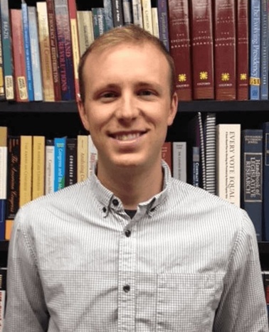 A smiling man wearing a gray checked shirt standing in front of a bookshelf filled with various books.