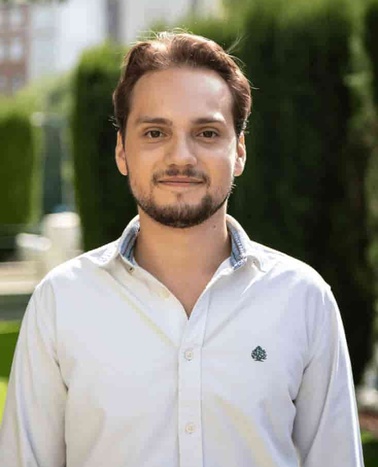 A portrait of a man with a beard standing outdoors with greenery in the background.