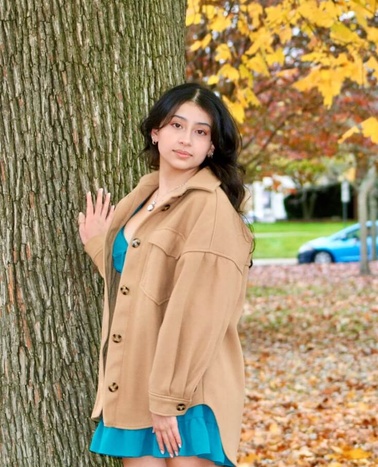 A young woman in a beige coat and blue skirt stands beside a tree with autumn leaves.