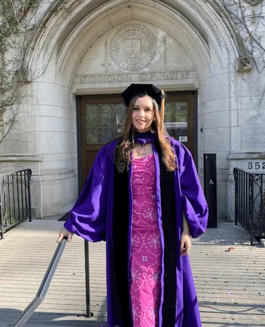 A young woman in a purple graduation gown and cap stands in front of a school entrance.