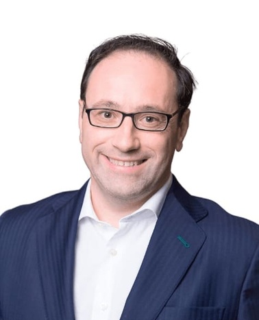 A professional portrait of a smiling man wearing glasses, a blue suit, and a white shirt.