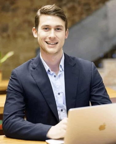 A young businessman smiling at the camera while working on his laptop in an office setting.