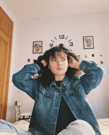 A young individual in a denim jacket is sitting in a bedroom with a simplistic clock design above their head.