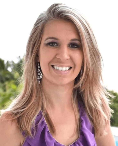 A smiling woman with long blonde hair, wearing a purple sleeveless top and decorative earrings, standing outdoors.