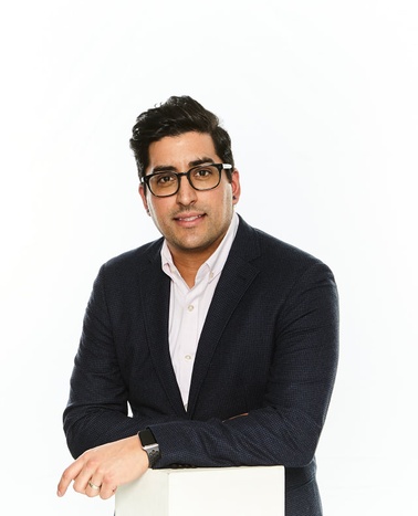 A man in a blazer and glasses leaning on a white podium against a white background.