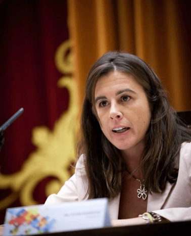 A woman is speaking at a formal event, seated with a microphone in front of her and a blurred ornate backdrop.