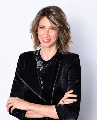 A professional woman with shoulder-length hair is smiling in a black blazer over a lace top, standing against a white background.