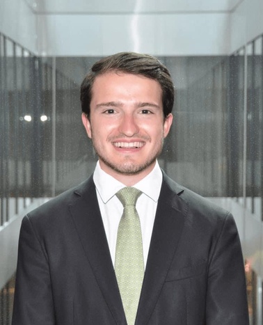 A young man in a dark suit with a light green tie smiling in a modern building interior.