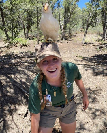A young woman smiling while a bird perches on her head in a natural setting.