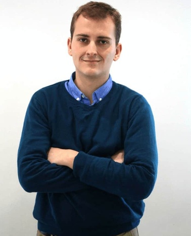 A young man in a blue sweater and blue shirt standing with his arms crossed and smiling against a white background.