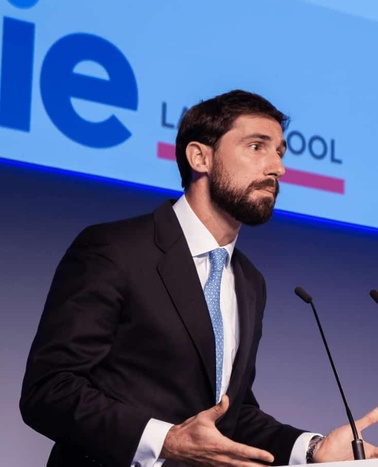 A man in a dark suit and blue tie speaking at a podium with a 'Le Cool' banner in the background.