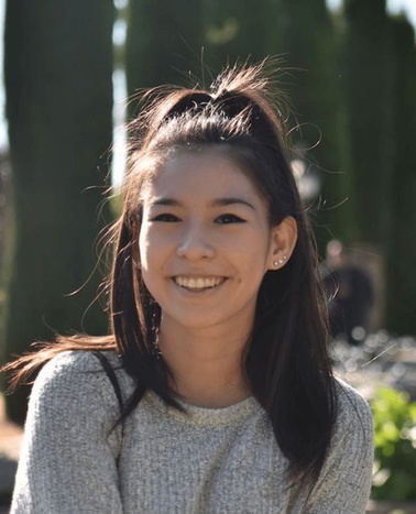 A young woman smiling in a park with trees in the background.