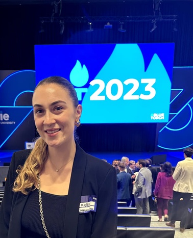 A woman in a business suit smiling at a graduation event, with '2023' displayed prominently in the background.