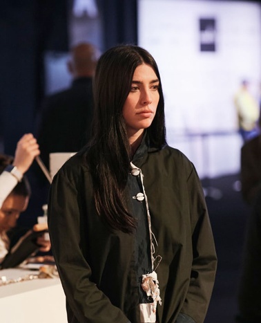 A woman with long black hair and a dark coat stands in a busy indoor setting, looking thoughtful.