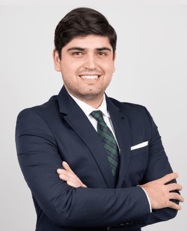 A smiling man in a navy blue suit with a white shirt and green striped tie standing with crossed arms against a grey background.