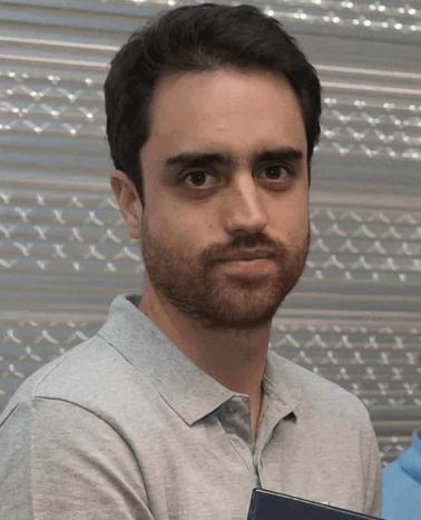 A man with a beard and short dark hair, wearing a grey shirt, standing indoors with a patterned backdrop.