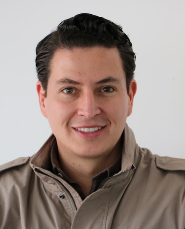Portrait of a smiling man with slicked-back hair wearing a beige jacket against a white background.