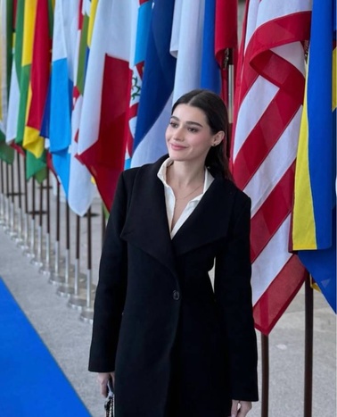 A woman wearing a black coat stands in front of international flags.
