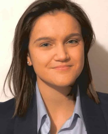 A young woman with dark hair wearing a dark suit and a light blue shirt smiling at the camera.
