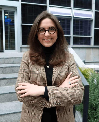 A woman in a plaid blazer and glasses smiling on steps outside a building.