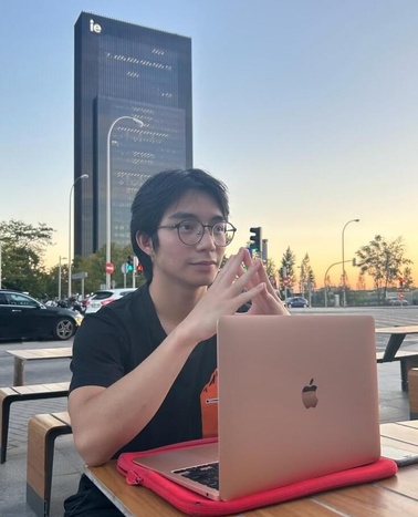 A young man is sitting at an outdoor table with a laptop, with a tall building in the background during sunset.