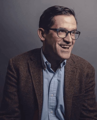 A portrait of a smiling man wearing glasses, a brown blazer, and a blue shirt.