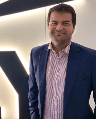 A man in a blue blazer and striped shirt standing in a modern interior with backlit large letter 'Y' on the wall.