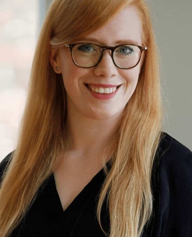 A portrait of a smiling woman with long red hair wearing glasses and a black blouse.