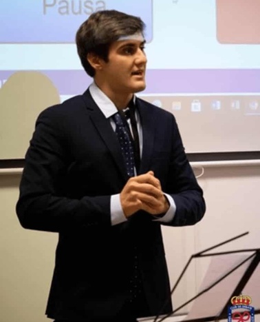 A man in a suit and tie giving a presentation in a classroom setting with a digital screen in the background showing the word 'Pausa'.