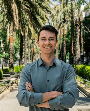 A smiling young man with arms crossed standing in a park with palm trees in the background.